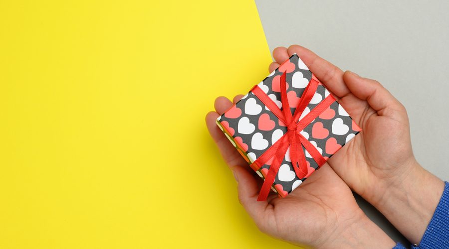 female hands hold cardboard box with red ribbon on yellow background, top view, copy space